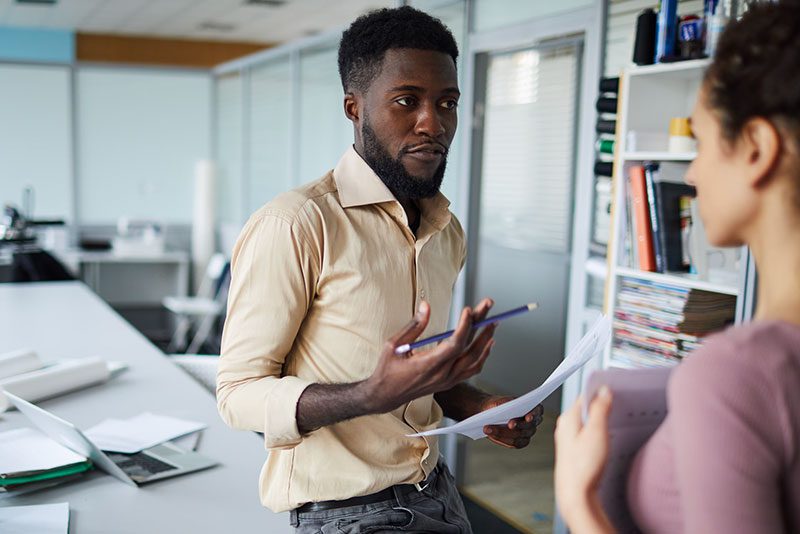 A man in a beige shirt holds a pen and papers, having a discussion with a woman in an office about CS Operations. He's leaning against a desk with a laptop, surrounded by shelves filled with books.