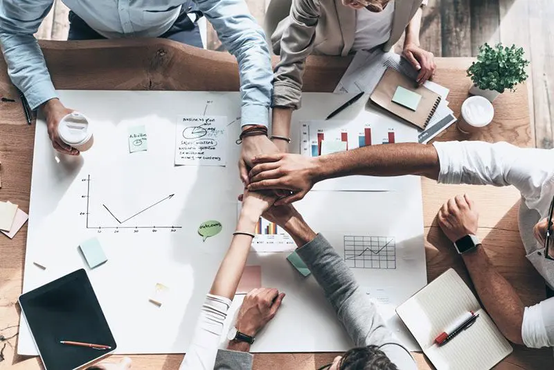 A group from the CS Ops Team gathers around a table with charts and papers, stacking their hands in unity. On the table are graphs, notebooks, a coffee cup, and sticky notes. A small plant sits in the corner, symbolizing the collaborative spirit of operations teamwork.