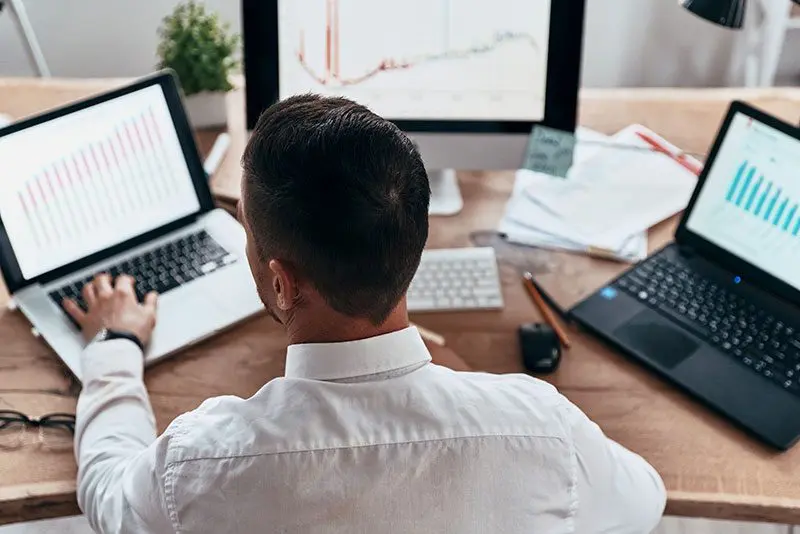 A person in a white shirt, part of the CS Ops Team, sits at a desk with two laptops and a desktop monitor displaying graphs. Papers, a plant, and office supplies clutter the wooden desk, reflecting the dynamic operations environment.