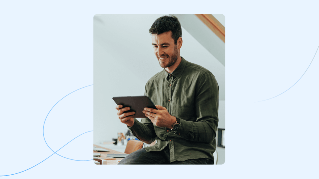 A person with short dark hair and a beard, wearing a green shirt, is smiling while looking at an AI tool displayed on their tablet. Seated in a bright room with a sloped ceiling, they appear engaged and thoughtful. The light blue background adds to the room's inviting atmosphere.